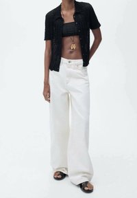Chemise noire à manches courtes boutonnée sur un crop top noir, associée à un pantalon blanc à jambes larges et des sandales noires, avec un design minimaliste.