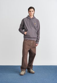 Grauer Kapuzenpullover mit Vorderer Tasche und rotem Logo, kombiniert mit braunen lässigen Hosen und beigen Stiefeln, vor einem hellen Hintergrund.