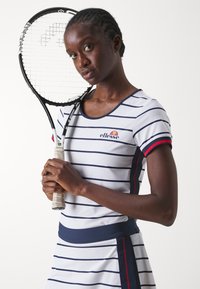Vestido blanco con rayas marinas, de manga corta y diseño ajustado. Presenta un escote redondo y acentos en rojo. La modelo sostiene una raqueta de tenis.