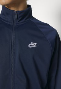 Námořnická modrá zipová bunda Nike s vyvýšeným límcem, vyrobená z hladké látky, s nápisem stříbrné barvy na levém prsu a kontrastními ramenními panely.