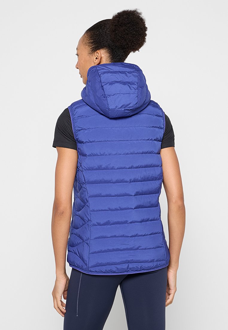 Frau trägt eine ärmellose, blaue Pufferweste mit Kapuze, ein schwarzes Kurzarmshirt und dunkle Leggings, steht mit dem Rücken zur Kamera vor einem weißen Hintergrund.