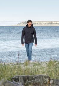 Mustat hupparitakki vetoketjupusseilla, yhdistettynä sinisiin farkkuihin ja vaaleisiin kenkiin; kävelee rantaa pitkin veden ja ruohikon lähellä.