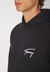 Hoodie in cotone nero con tasca frontale e logo "Tommy" ricamato in bianco sul petto sinistro. Il tessuto ha una texture liscia.