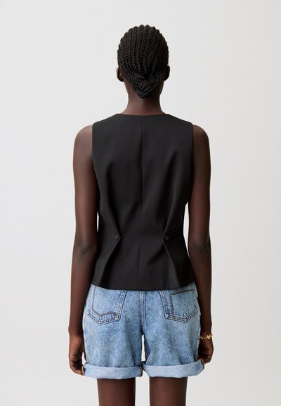 Top nero senza maniche con una forma aderente, caratterizzato da una vita definita e due bottoni sul retro; abbinato a pantaloncini di jeans azzurri arrotolati.