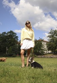 Gele sweatshirt, witte rucheskirt en zwarte sandalen. Een zwart-witte corgi zit ernaast op een grasveld onder een blauwe lucht met wolken.