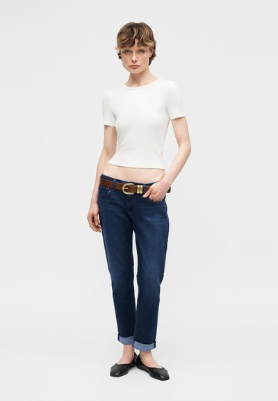 Νεαρό άτομο που φοράει εφαρμοστή λευκή κοντομάνικη μπλούζα, σκοτεινά τζιν με γυρισμένα μπατζάκια και καφέ ζώνη, καθώς και μαύρα επίπεδα παπούτσια, στέκεται μπροστά σε απλό φόντο.