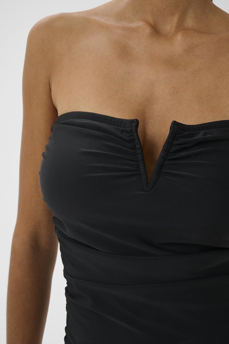 Frau trägt schwarzes trägerloses Top mit V-förmigem Ausschnitt, zeigt Schultern und oberen Brustbereich vor einfachem Hintergrund.