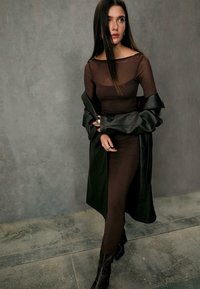 Braun eng anliegendes Kleid mit transparenten Einsätzen, darunter ein langer schwarzer Leder-Mantel, kombiniert mit strukturierten kniehohen Stiefeln. Glattes und elegantes Design.