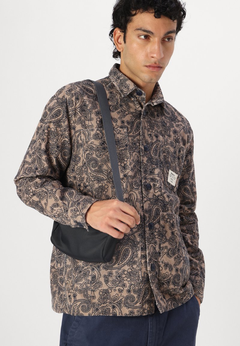 Chemise à manches longues avec boutons en brun clair ornée d'un motif paisley foncé, associée à un petit sac en bandoulière noir. Texture lisse et coupe décontractée.