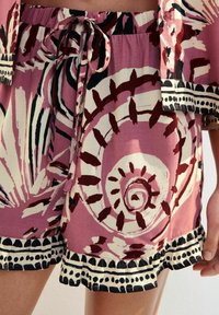 Kortbyxor i lätt tyg med en rosa bakgrund, djärva spiral- och blommönster i burgundy och svart, med volangkant i svart.