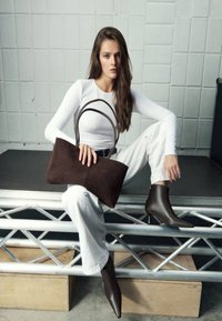 Brown suede handbag with dual handles, sleek design, and smooth texture. Worn with white fitted top and off-white pants, featuring pointed brown ankle boots.