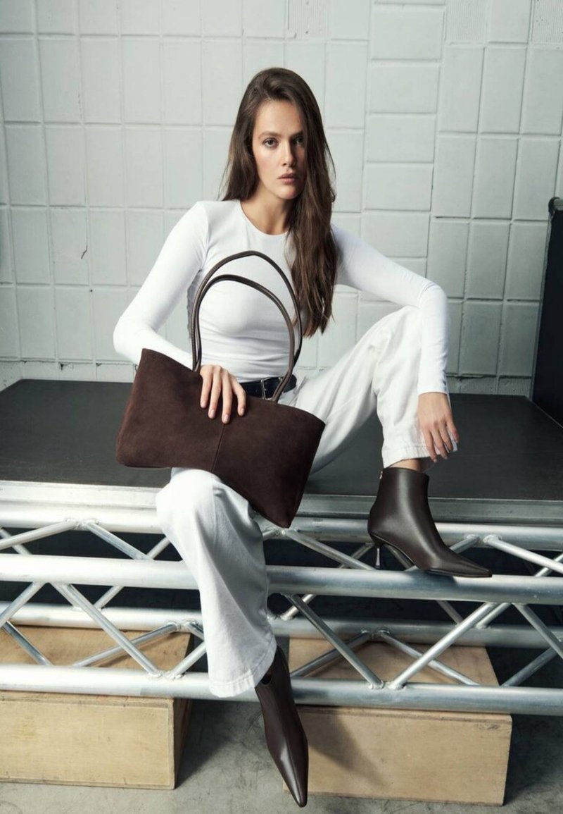 Brown suede handbag with dual handles, sleek design, and smooth texture. Worn with white fitted top and off-white pants, featuring pointed brown ankle boots.