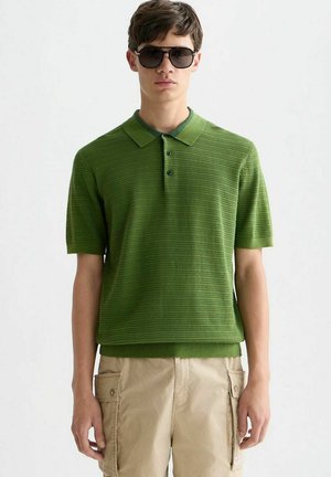 Junger Mann trägt grünes, strukturiertes kurzärmeliges Poloshirt, beige Cargoshorts und dunkle, übergroße Sonnenbrille und steht vor einem einfarbigen Hintergrund.