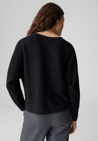 Schwarzer, strukturierter Pullover mit weitem Ausschnitt und langen Ärmeln, kombiniert mit grauen Hosen. Das Material wirkt weich und leicht gestrickt.