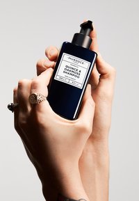 Dunkelblaue Flasche mit Murdock Quitte & Eichenmoos Shampoo mit einem schwarzen Pumpspender, versehen mit einem weißen Etikett mit schwarzem Text, in zwei Händen gehalten.