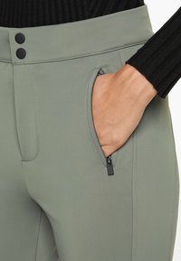 Hellgrüne Hose mit glatter Textur, ausgestattet mit einer Reißverschlusstasche und einem Knopfverschluss an der Taille. Teilweise sichtbares schwarzes Rippoberteil.