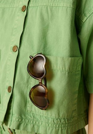 Heart-shaped brown sunglasses hanging from the front pocket of a light green button-up shirt.