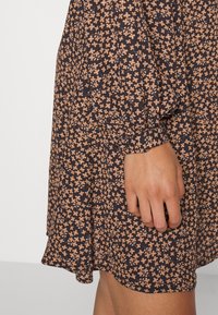 Svart klänning med ett blommönster i beige och brunt, med långa ärmar och en utställd fåll. Tyget verkar lätt och flödig.