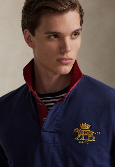 Μπλε ναυτικό πόλο με κόκκινο γιακά, διακοσμημένο με κεντητό χρυσό λιοντάρι με κορώνα. Ριγωτό εσώρουχο εμφανές στον γιακά.