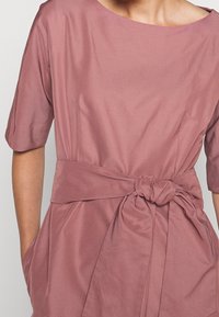 Růžové šaty z hladké tkaniny, s kulatým výstřihem, krátkými rukávy a detailem vázané pásky v pase se bočními kapsami.
