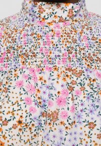 Blouse col haut avec empiècement froncé présentant un motif floral composé de fleurs roses, orange, marron et violettes sur fond blanc.