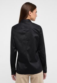 Schwarzes Langarmshirt aus glattem Stoff, mit klassischem Kragen, Knopfmanschetten und tailliertem Schnitt mit gerader Rückennaht.