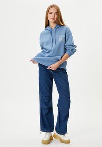 Sudadera azul clara con un bolsillo frontal y capucha con cordones; combinada con pantalones de mezclilla oscuros. El modelo lleva zapatos blancos voluminosos.