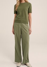 Woman wearing textured olive green short-sleeve top, matching wide-leg trousers, and cream slip-on shoes against a plain background.