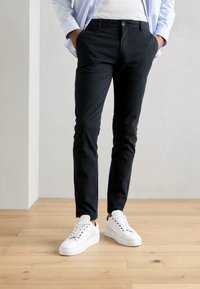 Zwarte slim-fit chinos met zijzakken, een knoopsluiting, gedragen met witte sneakers, tegen een lichte houten vloer en een neutrale muur.