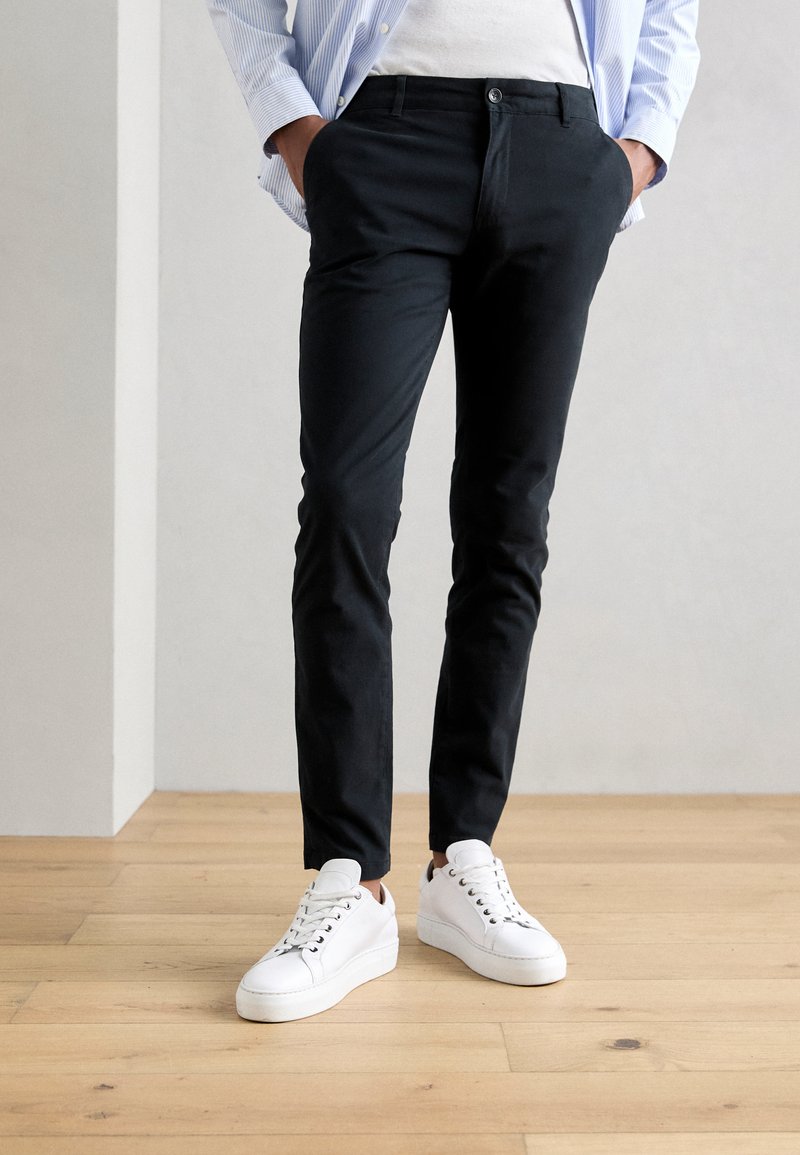 Zwarte slim-fit chinos met zijzakken, een knoopsluiting, gedragen met witte sneakers, tegen een lichte houten vloer en een neutrale muur.