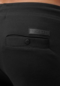 Nahaufnahme von schwarzen Stoffhosen mit einer Gesäßtasche, die einen Knopf und ein rechteckiges Patch mit der Aufschrift "INDICODE" zeigt.