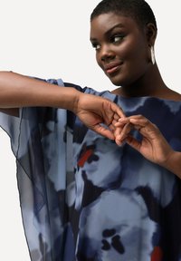 Blusa azul fluida y transparente con un patrón floral en tonos azul oscuro y acentos rojos. Presenta un escote amplio y mangas sueltas.