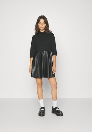 Pull noir à manches trois-quarts associé à une jupe noire en simili cuir. Le mannequin porte des chaussures à plateforme noires et des chaussettes blanches à la cheville.