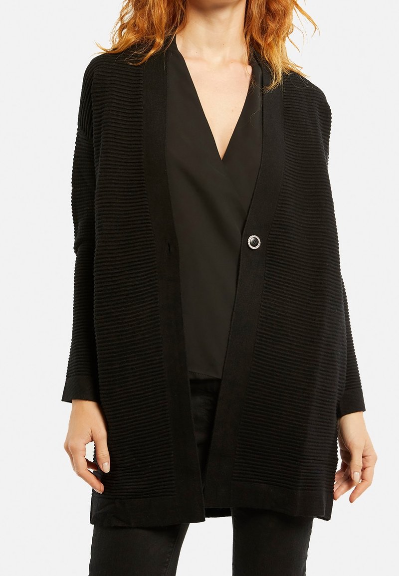 Černý kardigan, ribovaná textura, otevřený přední část s jedním knoflíkem, dlouhé rukávy, do délky kolen, kombinovaný s černým topem pod ním.