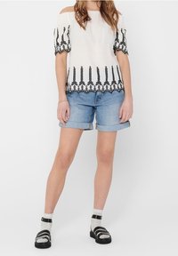 Haut blanc décolleté avec broderie noire et ourlet festonné, associé à un short en jean retroussé et des sandales noires portées avec des chaussettes blanches.