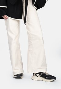 Jeans évasés blancs en tissu coton associés à des baskets noires et beiges avec des accents en maille et en caoutchouc. Fond uni pour accentuer l'objet.