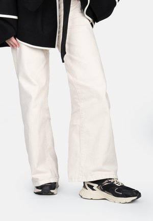 Jeans évasés blancs en tissu coton associés à des baskets noires et beiges avec des accents en maille et en caoutchouc. Fond uni pour accentuer l'objet.