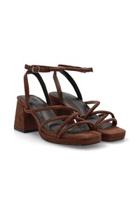 Tacones de ante marrón con tiras, tacón cuadrado, diseño de tiras cruzadas y suela texturizada, con una correa en el tobillo que presenta un detalle de hebilla.