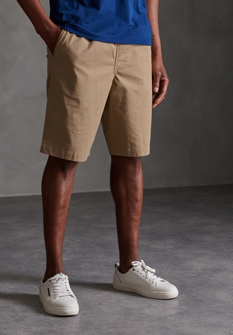Homme portant un short beige jusqu'aux genoux, une chemise bleue et des baskets blanches, debout sur un sol gris, les mains dans les poches.