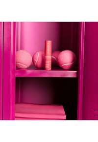 Rosa Schließfachregal mit vier rosa Tennisbällen, einem roten NUXE Prodigieuse Hyalu Augenstift und gefalteter rosa Stoff darunter.