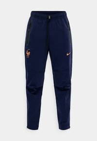 Στενό παντελόνι αθλητικού τύπου σε ναυτικό μπλε, με ελαστική μέση, πλαϊνές τσέπες με φερμουάρ, πορτοκαλί λογότυπο Nike Swoosh και το έμβλημα της Γαλλικής Ποδοσφαιρικής Ομοσπονδίας με δύο αστέρια.