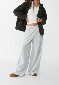 Tmavě šedá fleecová bunda se zipem vpředu, doplněná o volné světle šedé tepláky. Outfit doplňují hnědé boty s hrubou podešví.