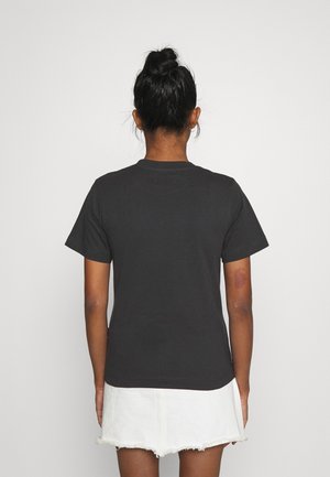 Vrouw met donker haar in een knot, gehuld in een zwart shirt met korte mouwen en een witte rafelige rok, met haar gezicht naar een effen lichtgrijze achtergrond.