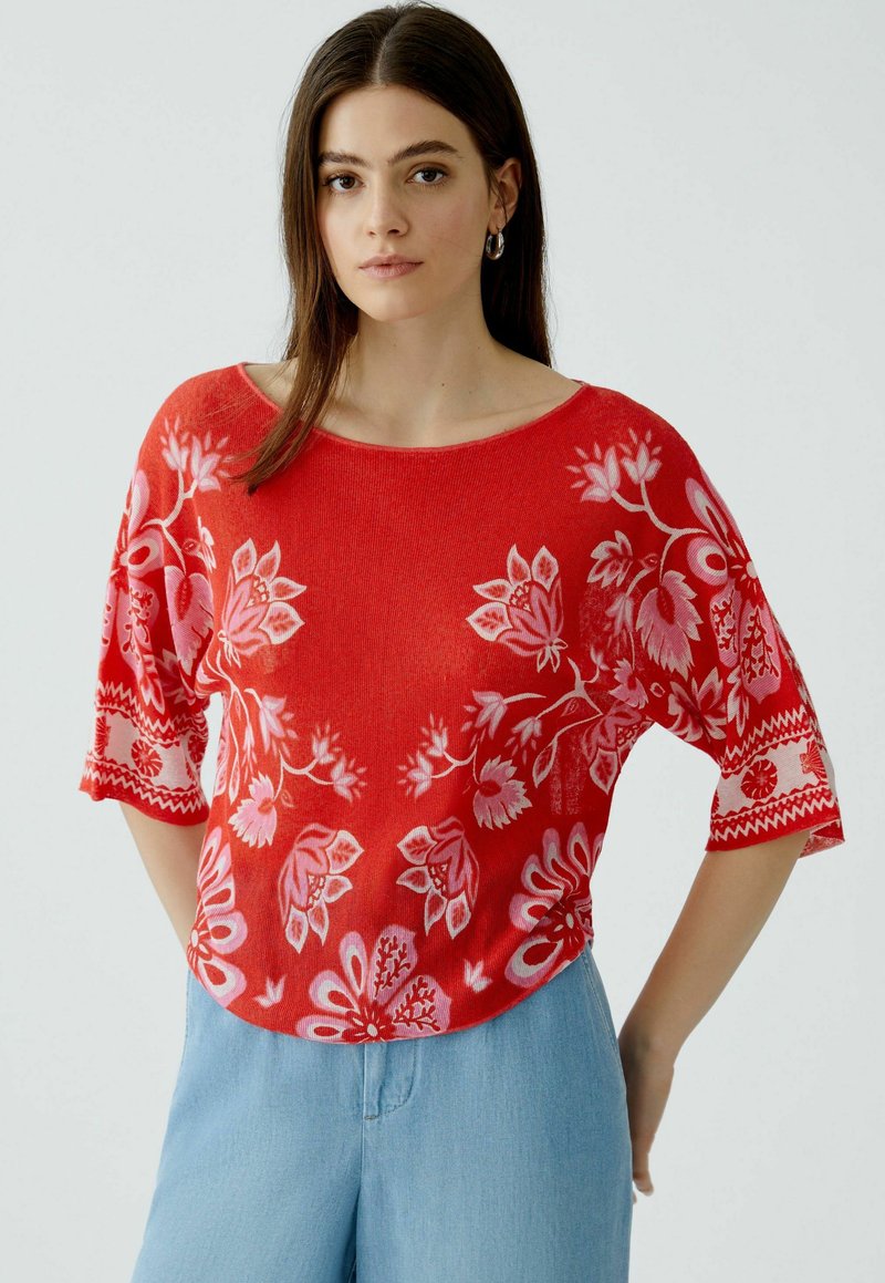 Rotes Strickoberteil mit floralen Mustern in Pink. Es hat einen weiten Ausschnitt und kurze Ärmel, mit einem geraden Saum und strukturiertem Stoff.