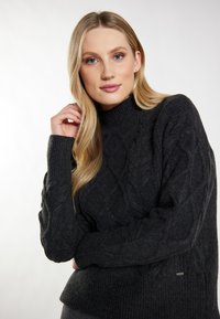 Mørkegrå kabelstrikket sweater med en høj ribbet halsudskæring, der har markante teksturerede mønstre og et lille logo-detalje ved kanten.