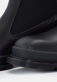 Gros plan sur des bottines Chelsea en cuir noir avec panneaux latéraux élastiques et semelles en caoutchouc texturées sur une surface blanche.
