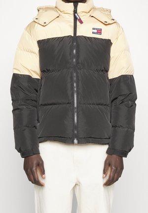 Chaqueta tipo acolchado con cierre de cremallera, que presenta una sección superior en crema y una sección inferior en negro, un parche de logo prominente y una capucha desmontable.