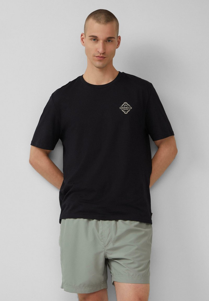Zwarte katoenen t-shirt met korte mouwen en een logo in goud, gecombineerd met lichtgroene shorts. Het ontwerp is eenvoudig en casual.
