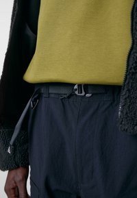 Olivgrüner Pullover, der unter einer schwarzen Jacke getragen wird, kombiniert mit dunkelblauen Hosen, die einen strukturierten Gürtel und einen verstellbaren Kordelzug haben.