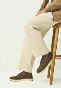 Brune suede loafers med en hvid gummisål, detaljeret syning og en lav profil; parret med lyse beige tapered bukser.